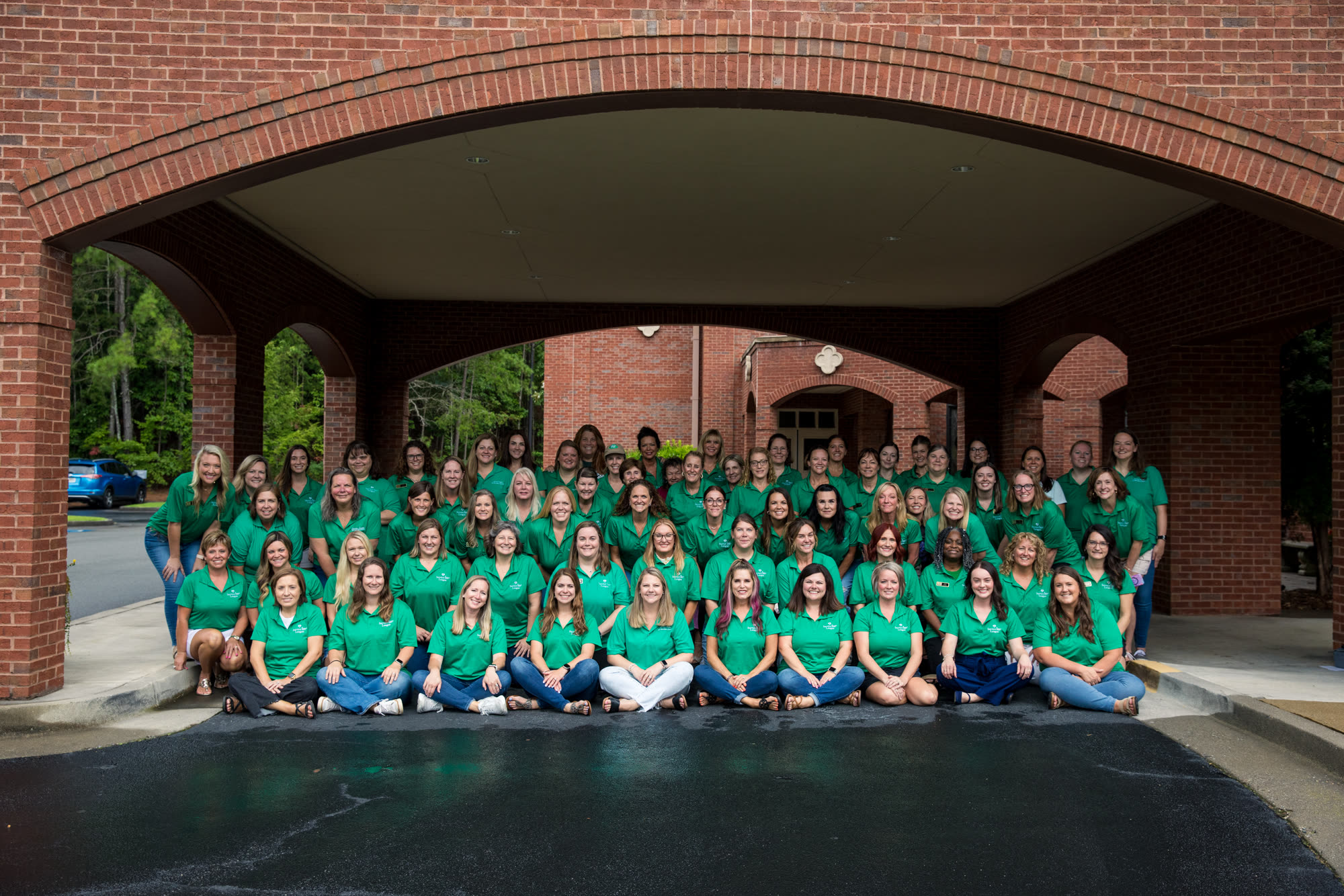 The 2025-26 Service League members, about seventy women in green league polos, gathered under the brick archway.