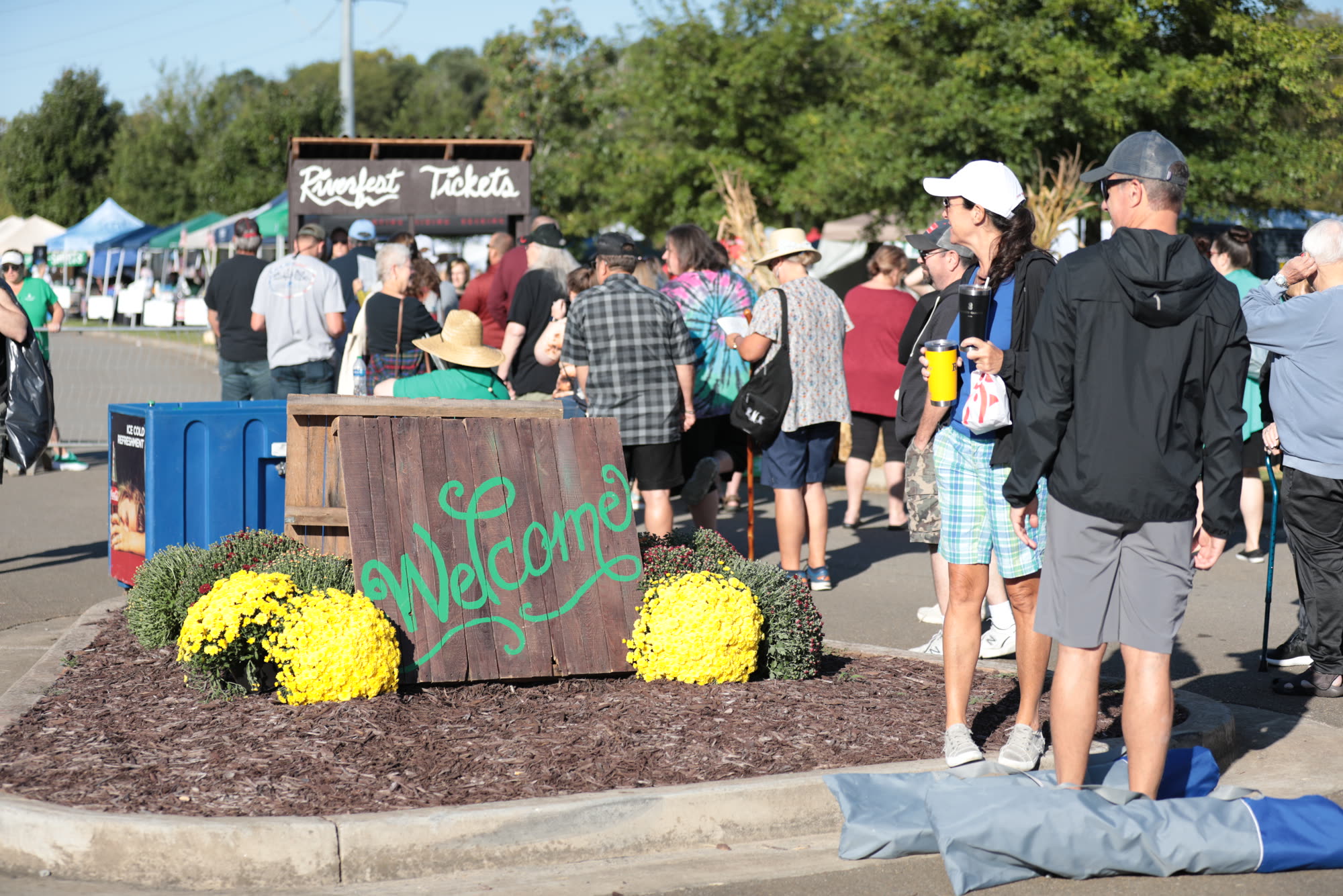 Festival-goers stream past a hand-painted Welcome sign ringed with yellow mums at the Riverfest front gate.